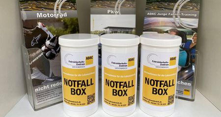 Drei weiße Plastikdosen mit Deckel, gebrandet im ADAC Design und mit der Aufschrift Notfallbox versehen, stehen nebeneinander. Im Hintergrund sind Flyer zu den Trainingsthemen Pkw-, Motorrad- und Pkw Junge-Fahrer-Training in Acrylspendern zu sehen. 