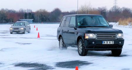 Im Vordergrund ein dunkler Jeep beim Durchfahren eines Slalom-Parcours, markiert durch mehrere Pylone. Im Hintergrund ein silberner Pkw ebenfalls in Slalomfahrt. Der Asphalt ist nahezu vollständig mit einer Schneeschicht bedeckt.