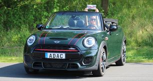 Gutschein für ein Pkw Intensiv-Training - Junge Frau mit blonden Haaren und Sonnenbrille in einem sommerlichen schwarzen Top; sie sitzt lächelnd im offenen dem dunkelgrünen Mini Cabrio. Mit beiden Händen streckt sie den Gutschein hoch aus mit geöffneten Dach.