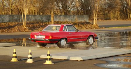 Ein roter Mercedes-Oldtimer mit H-Kennzeichen fährt von rechts über die Hydraulikplatte. Die Teststrecke ist bewässert. Ansonsten scheint es sich um einen sonnigen Herbsttag zu handeln.