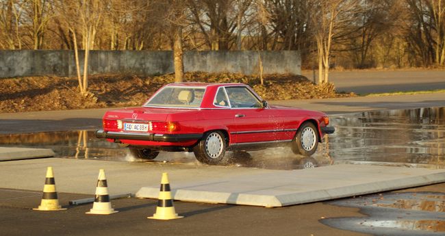 Ein roter Mercedes-Oldtimer mit H-Kennzeichen fährt von rechts über die Hydraulikplatte. Die Teststrecke ist bewässert. Ansonsten scheint es sich um einen sonnigen Herbsttag zu handeln.