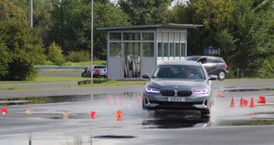 Der Fahrer lenkt seinen grauen BMW durch das Pylonentor. Die Scheinwerfer sind an, die Fahrbahn ist nass. Im Hintergrund stehen einige Bäume.