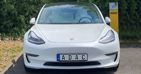Front-Aufnahme eines weißen E-Fahrzeugs der Marke Tesla. Im Hintergrund die gelbe, mit ADAC-Logo versehene Ladesäule mit eingestecktem Ladekabel.