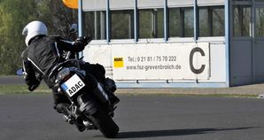 Motorrad Firmengruppen – Ein Motorradfahrer in Schutzkleidung in Kurvenlage auf trockenem Asphalt. Im Hintergrund ist Modulhaus C zu sehen.
